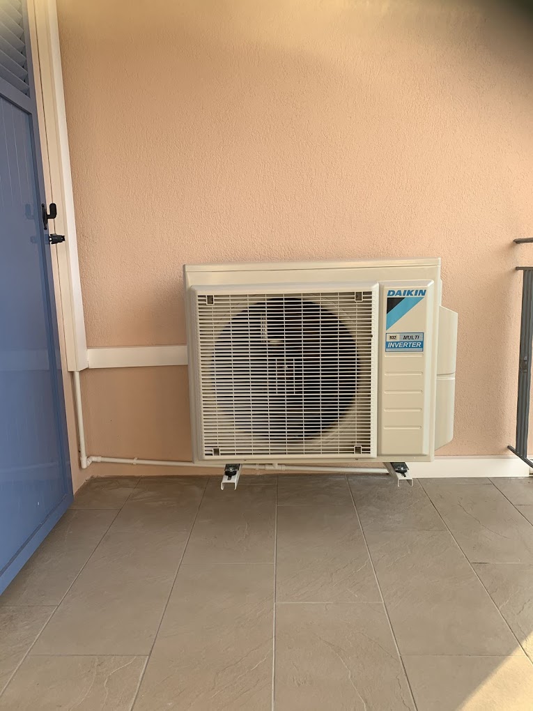 Unité de climatisation Daikin extérieure installée sur un balcon beige avec porte bleue.